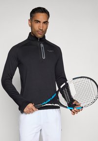 Pulover sport negru cu fermoar și guler înalt, fabricat dintr-un material elastic. Modelul ține o rachetă de tenis cu un design albastru și negru, purtând pantaloni scurți albi.