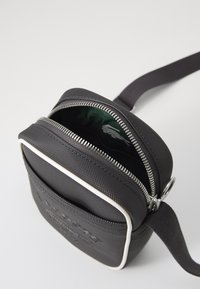 Black fabric crossbody bag with a textured surface, silver zipper, and white trim. Includes an interior pocket with a green patterned lining.