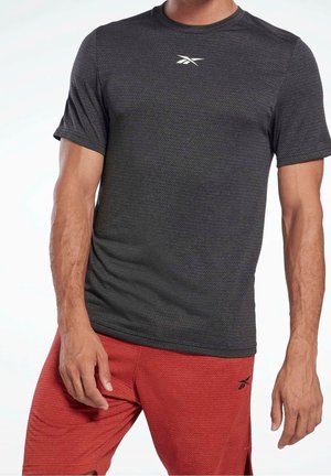 Mann trägt ein eng anliegendes schwarzes, strukturiertes Kurzarmhemd und rote, strukturierte Shorts, beide mit einem kleinen weißen Logo auf Brust und Oberschenkel.
