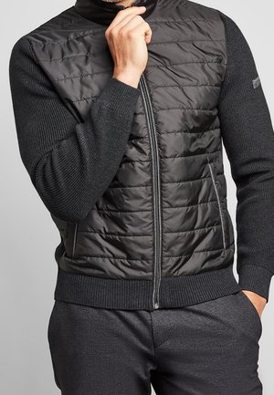 Homme portant un gilet matelassé noir à fermeture éclair avec manches et col en tricot, une main près du menton et l'autre dans la poche.