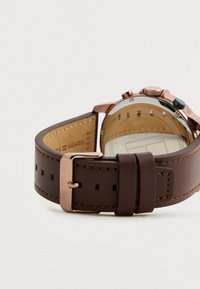 Brown leather watch strap with rose gold buckle resting on a white surface, showing the back of the watch case and strap detail.