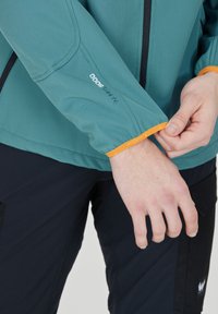 Teal vattentät jacka med orangea detaljer på ärmarna, som visar elastiskt material och en figurnära design. Logotypen syns på ärmen.