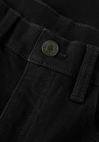 Gros plan sur un jean en denim noir présentant un bouton en métal avec un texte gravé, des coutures contrastées et une texture lisse.