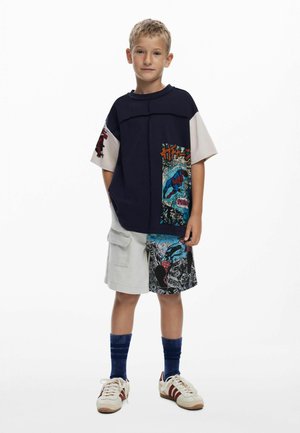 Young boy standing with hands in pockets wearing navy and white Spider-Man graphic T-shirt, mismatched shorts, blue socks, and white sneakers.