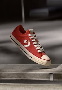 Sneaker di tela rossa con logo a stella bianca, punta in gomma crema e lacci testurizzati. Presenta accenti blu sulla suola e un design a basso profilo.