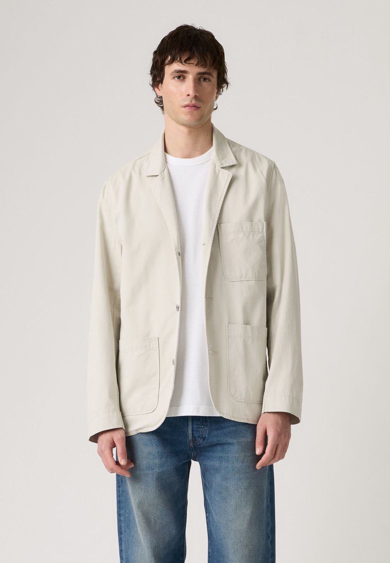 Homme portant une veste boutonnée beige clair sur un t-shirt blanc et un jean bleu, debout devant un fond clair uni.