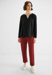 Maglietta nera a maniche lunghe con scollo a V e bottoni, abbinata a pantaloni dritti bordeaux e sneakers bianche su uno sfondo semplice.
