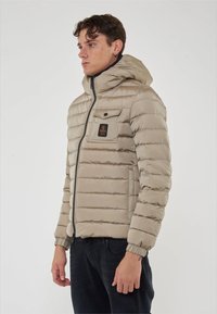 Giacca trapuntata beige con cuciture orizzontali, cappuccio fissato, zip frontale e tasca sul petto con logo. Texture liscia e lucida.