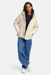 Veste crème matelassée avec capuche, dotée d'une fermeture éclair à l'avant, d'un design matelassé et de poches latérales. Associée à un jean bleu à jambes larges et à des baskets blanches.