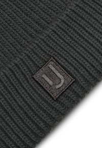 Har grey beanie-lue som er strikket med strukturert ribbestrikk og har en firkantet svart etikett med en hvit logo. Materialet ser mykt og varmt ut.
