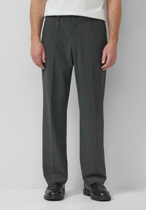 Man draagt donkergrijze nette broek en zwarte schoenen, staand tegen een effen lichtgrijze achtergrond.