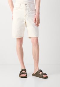 Shorts en denim blanc avec une coupe droite et des ourlets retroussés, accompagnés de sandales plates marron à brides réglables et semelles texturées.