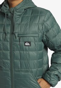 Green quilted jacket with a hood, featuring a zippered chest pocket and elastic cuffs. Made of smooth, lightweight material.