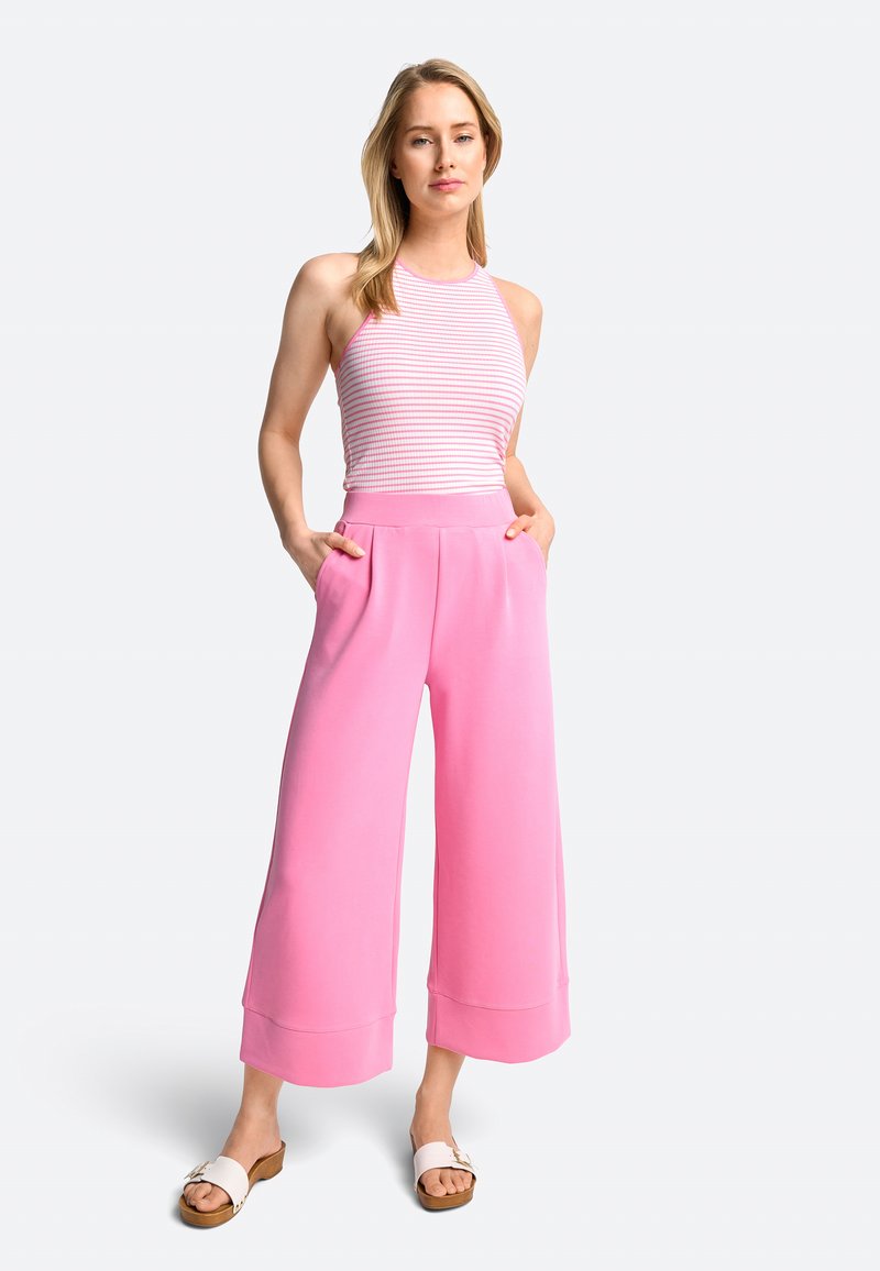 Roze wijde broek van gladde stof, gecombineerd met een gestreept mouwloos topje. Model staat met handen in de zakken, draagt lichte sandalen.