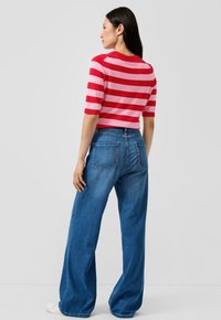 Vrouw in een rood- en roze gestreept top met korte mouwen en een high-waisted wijd uitlopende blauwe spijkerbroek, staand tegen een effen achtergrond.