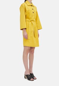 Abito camicia giallo con colletto, frontale con bottoni e vita con legatura. Presenta maniche lunghe e una vestibilità rilassata, abbinato a mule nere con tacco.