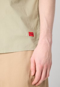 Camisa de algodón verde claro con una etiqueta de logotipo rectangular roja. Textura suave, el dobladillo se sitúa por encima de la tela marrón claro. Mano descansando cerca.