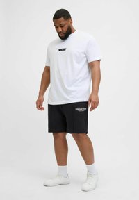 T-shirt en coton blanc avec logo noir, associé à un short noir portant le texte "Originals Studio", porté avec des baskets blanches.