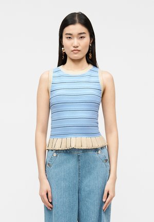 Kvinne med langt mørkt hår iført en ermeløs blå og beige stripete strikkegenser og høytlivs lysblå jeans, stående mot en hvit bakgrunn.