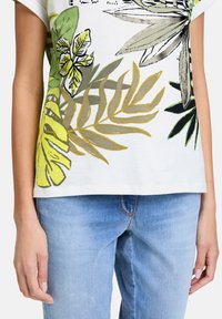 Biała bawełniana koszulka z zielonymi i żółtymi nadrukami liści; krótki rękaw i luźny krój. Połączona z jasnoniebieskimi dżinsami.
