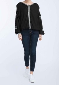 Blusa nera a maniche lunghe con scollatura a V e ricami bianchi decorativi lungo la parte frontale e le maniche, abbinata a jeans scuri e sneakers bianche.