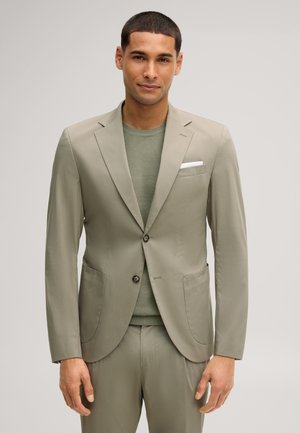 Homme portant une veste de costume beige clair par-dessus un pull en maille verte, sur un fond uni clair.