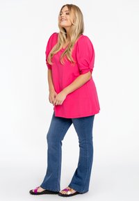 Heldere roze top met korte mouwen en geplooide details, gecombineerd met blauwe flared jeans. Het model draagt zwarte sandalen met meerkleurige banden.