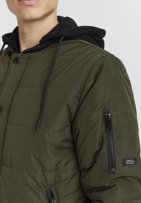 Giacca trapuntata verde oliva con cappuccio in pile nero, chiusure a scatto e zip, e tasche laterali; presenta un'etichetta a contrasto sulla manica.