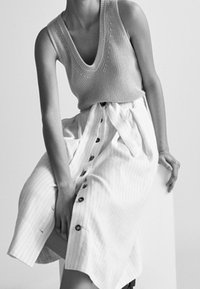 Femme portant un top en maille sans manches et une jupe rayée boutonnée et nouée à la taille, assise sur un bloc blanc sur un fond uni.