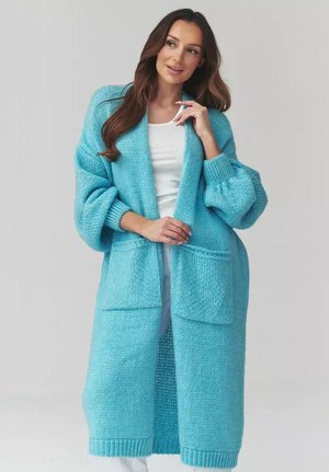 Kobieta z długimi brązowymi włosami, ubrana w długi, oversize'owy, jasnoniebieski sweter na drutach, nałożony na biały top i spodnie, stoi na szarym tle.