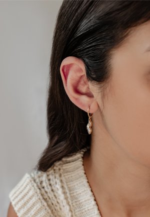 Gros plan sur l'oreille d'une femme aux cheveux foncés portant une boucle d'oreille pendante en or avec trois pierres claires, sur un fond neutre flou.