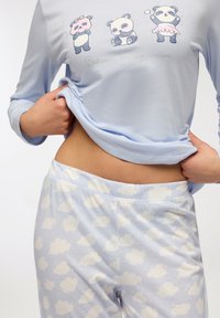 Top in morbido azzurro con grafiche di panda e la scritta "Credi nella routine di bellezza", abbinato a pantaloni azzurri chiari con motivi di nuvole bianche.