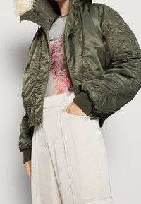 Chaqueta bomber verde oliva con capucha forrada de piel sintética, bolsillo con cremallera en la manga y puños acanalados, combinada con pantalones de pierna ancha de color claro.