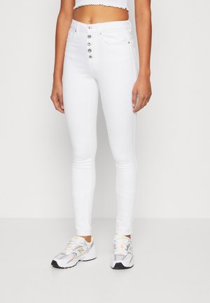 Kobieta ubrana w białe, obcisłe dżinsy z wysokim stanem z srebrnymi guzikami oraz białe i szare sportowe sneakersy, stojąca na jednolitym tle.