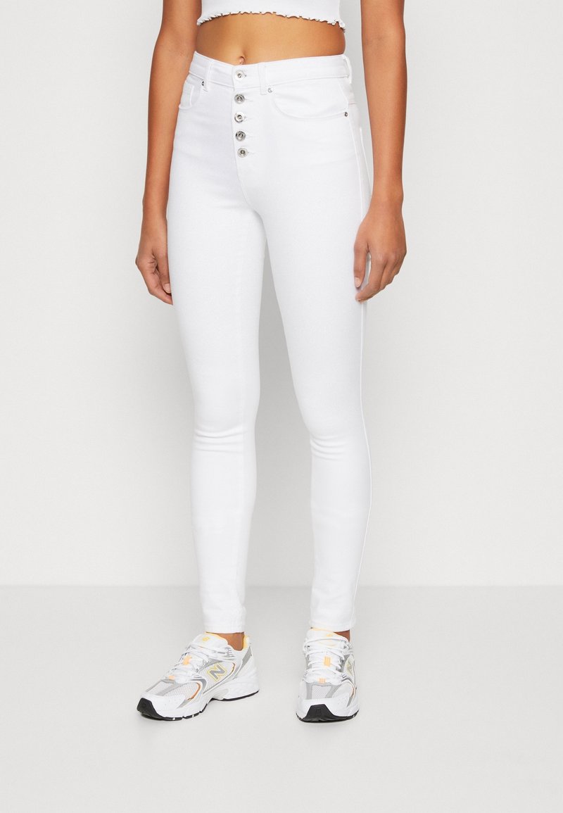 Femme portant un jean skinny blanc taille haute avec des boutons argentés et des baskets blanches et grises, debout devant un fond uni.