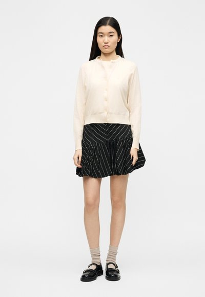 Γυναίκα που φοράει μπεζ ζακέτα με κουμπιά, μαύρη φούστα με λευκές διαγώνιες ρίγες, μπεζ κάλτσες και μαύρα παπούτσια με αγκράφα, στέκεται μπροστά σε λευκό φόντο.