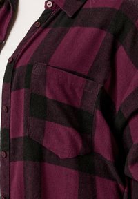 Camicia in flanella a quadri bordeaux e nera con design a bottoni, taschino sul petto e texture morbida. Presenta un colletto arrotondato e bottoni scuri.