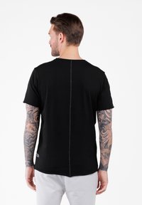 Mężczyzna z tatuażami na rękach, ubrany w czarną koszulkę z pionowym szwem na plecach i jasne spodnie, odwrócony plecami do kamery.