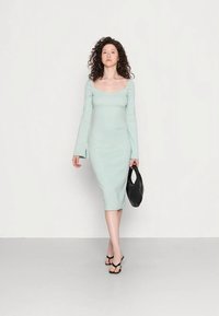 Mulher a caminhar para a frente, vestindo um vestido midi justo de tom verde claro, sandálias pretas, brincos de argola dourados e a segurar uma bolsa preta.