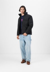 Sort pufferjakke med høj krave, lynlåslukning og firkantet quiltet mønster, som er parret med lyseblå jeans og brune sko.
