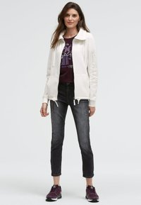 Weißer Zip-Hoodie mit Kordelzügen und Seitentaschen, kombiniert mit einem dunkelvioletten Grafik-T-Shirt, dazu dunkelgraue Jeans und lila Sneakers.