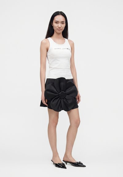 Femme portant un haut blanc sans manches et une jupe noire avec un grand détail de fleur en tissu, associée à des chaussures slingback noires à bout pointu.