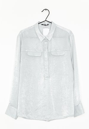 Blusa gris claro de manga larga con cuello, dos bolsillos de solapa en el pecho y botones ocultos, colgada en una percha negra sobre un fondo blanco.