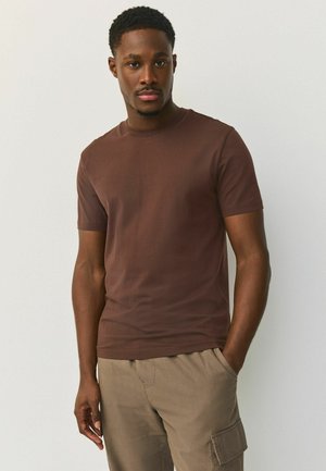 Mężczyzna w brązowej koszulce z krótkim rękawem i beżowych spodniach typu cargo stoi z jedną ręką w kieszeni na tle jednolitego jasnego tła.