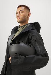 Sort vandtæt jakke med hætte, med en lynlåslomme og tekstureret overflade. Modellen holder en mat sort hjelm. Grøn bundlag er synligt.