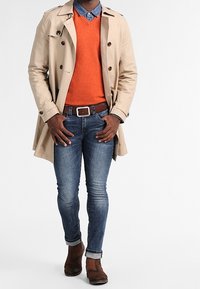 Beige trenchcoat över en orange tröja och denimskjorta, kombinerat med blåjeans och bruna skor. Noterbara detaljer inkluderar uppvikta ärmar.