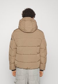 Giubbotto puffo marrone con cappuccio, design trapuntato e polsini in elastico. Il tessuto ha una texture morbida, con una vestibilità comoda e senza loghi visibili.
