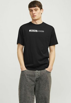 Zwarte katoenen T-shirt met korte mouwen, voorzien van een wit "JACK & JONES" logo aan de voorkant. Draagt met lichtgrijze spijkerbroek.