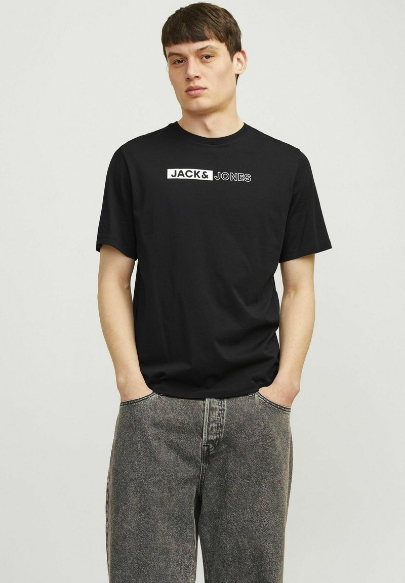 Zwarte katoenen T-shirt met korte mouwen, voorzien van een wit "JACK & JONES" logo aan de voorkant. Draagt met lichtgrijze spijkerbroek.
