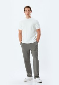 Vit kortärmad skjorta, grå texturerade byxor och vita sneakers. Modellen står med händerna i fickorna, neutral bakgrund.
